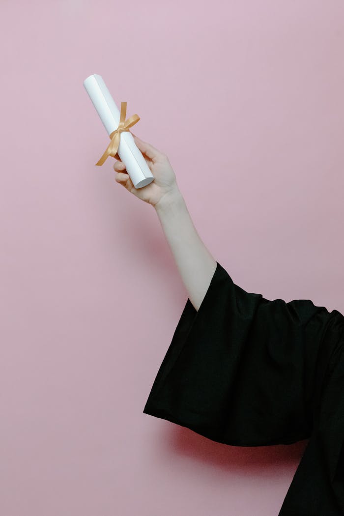 Hand of a graduate wearing a gown holding a diploma with a pink background.